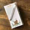 The To Moo Heelan Coo List Pad showcases the phrase To moo... at the top, adorned with an enchanting illustration of Moo Heelan Coo surrounded by flowers at the bottom. This delightful list pad includes a pen and is beautifully presented on a rustic wooden surface, making it an ideal stationery choice for cow lovers and rustic home decor enthusiasts.