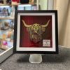 Discover our stunning framed artwork featuring a Highland cow, masterfully crafted with textured layers in vibrant green and luxurious gold hues against a striking red plaid background. This captivating piece is elegantly displayed on a stand in a retail store setting, surrounded by an array of items on neatly organized shelves. Perfect for art lovers and home decorators seeking unique statement pieces.