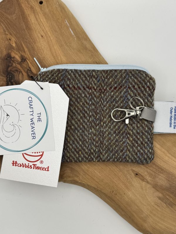 A compact Harris Tweed Clippy Purse, featuring a convenient zipper and durable metal clip, rests elegantly on a wooden surface. Prominent tags showcase The Crafty Weaver branding alongside the genuine Harris Tweed label, emphasizing its classic woven design.