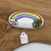A beautifully painted rock on a wooden surface showcases a vibrant rainbow design. On the left side of the rock, the phrase Good Luck is artfully inscribed within a cloud. The right side features an eye-catching pot of gold adorned with a shamrock. This unique item includes an attached price tag with handwritten numbers, making it a perfect addition to any decor collection. Ideal for those seeking good fortune and colorful charm in their home decor piece!.