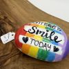 Vibrant Painted Rock with Inspiring Message: Dont Forget to Smile Today Decorated with Rainbow Stripes and Black Hearts on a Wooden Surface. Available for $8.00.