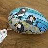 A hand-painted stone featuring puffin art showcases two vividly colored puffins with orange beaks. These charming seabirds are perched on realistic gray rocks, set against a striking blue background. A number tag attached via a string enhances its uniqueness. This beautiful decorative stone sits elegantly on a wooden surface, perfect as unique coastal decor or a nature-inspired gift for bird lovers.
