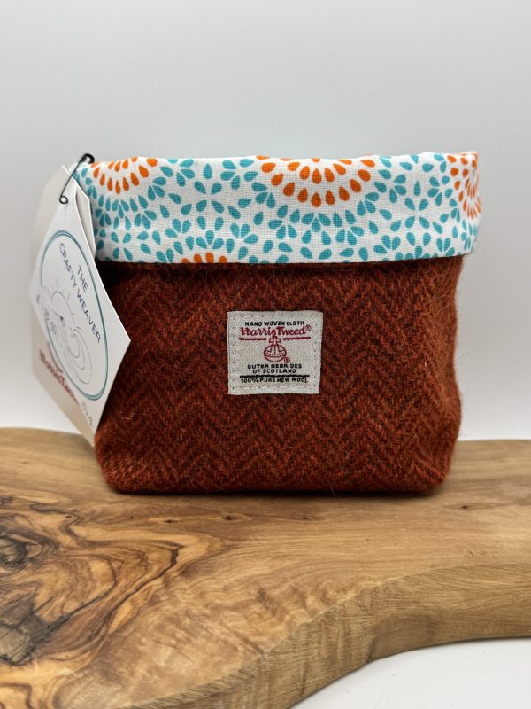 A charming, handcrafted fabric bag rests on a wooden surface. This unique bag showcases a rust-colored herringbone pattern complemented by a white top edge adorned with an eye-catching blue and orange floral design. Both a label and a hanging tag are affixed to the bag for added detail.