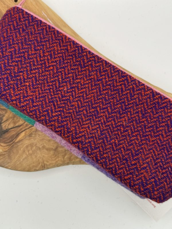 A rectangular woolen fabric featuring a red and purple herringbone pattern is elegantly draped over a wooden surface, enhancing its visual appeal. The intricate weave creates an eye-catching, textured appearance that adds depth to the fabrics design.