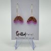 Gold hoop earrings featuring pink and gold glitter heart-shaped pendants are showcased on a card displaying the Spotted Petal branding. The shimmering heart pendant earrings rest elegantly on a transparent stand against a soft, light background.
