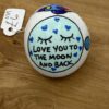 A beautifully painted rock on a rustic wooden surface showcases a serene blue moon with closed eyes, encircled by charming blue hearts. The inscription reads LOVE YOU TO THE MOON AND BACK. Attached to the rock is an affordable price tag displaying 1.99.