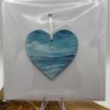 Introducing a heart-shaped ornament, meticulously hand-painted to showcase a tranquil seascape with serene blue skies and gentle waves. This exquisite keepsake evokes tranquility and makes an ideal gift for cherished memories. Perfectly displayed against a white background in clear packaging, it rests on a small acrylic easel atop a wooden surface, mirroring the elegance of a Keepsake Card. Perfect for those seeking unique gifts, coastal decor enthusiasts will love this ocean-inspired piece that combines artistry with sentimentality.