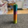 A vibrant spool of thread rests on a gray surface, showcasing lively colors that mirror the 3D Printed Telescopic Swords hilt. This colorful spool transitions through shades of green, blue, purple, pink, and orange, nestled in a softly blurred indoor setting with various objects.