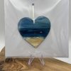 A hand-painted heart beautifully depicts a serene beach scene with two bathers enjoying the water. This charming artwork, elegantly showcased in clear packaging, rests gracefully on a wooden surface accompanied by an acrylic stand—ideal as a keepsake card or unique gift.