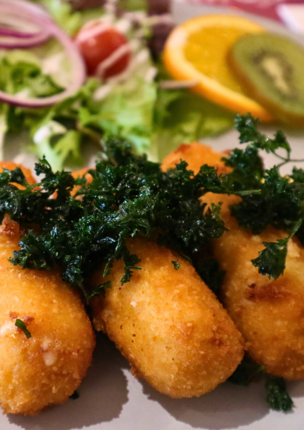 Drie garnaalkroketten op een rij, erbovenop ligt gefrituurde peterselie, geserveerd met een partje citroen en een slaatje.