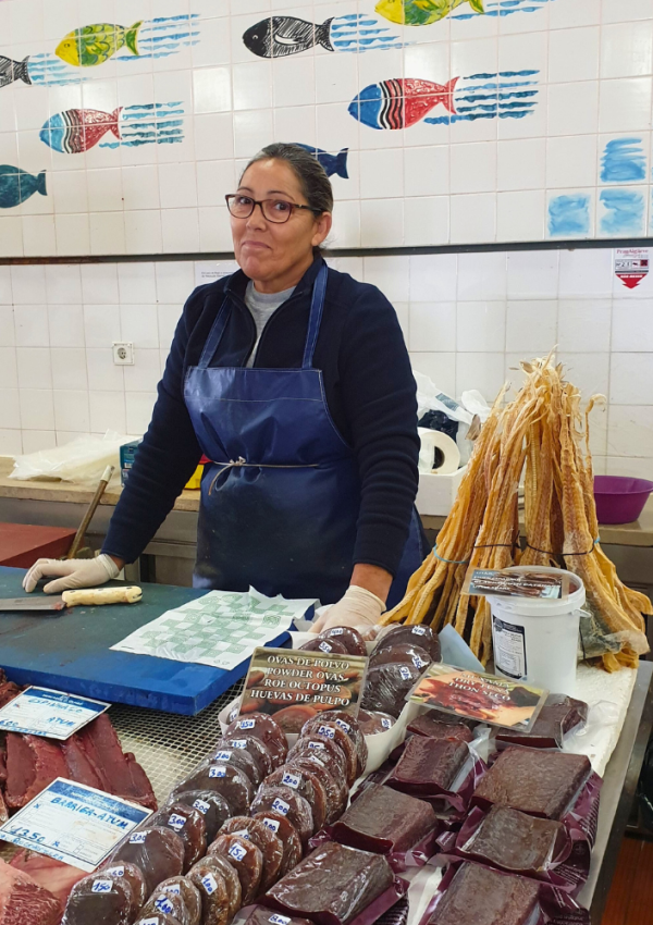 Een verkoopster in de markthal van Olhão in de Algarve bij haar koopwaar. Op de tegels achter haar staan vissen getekend.