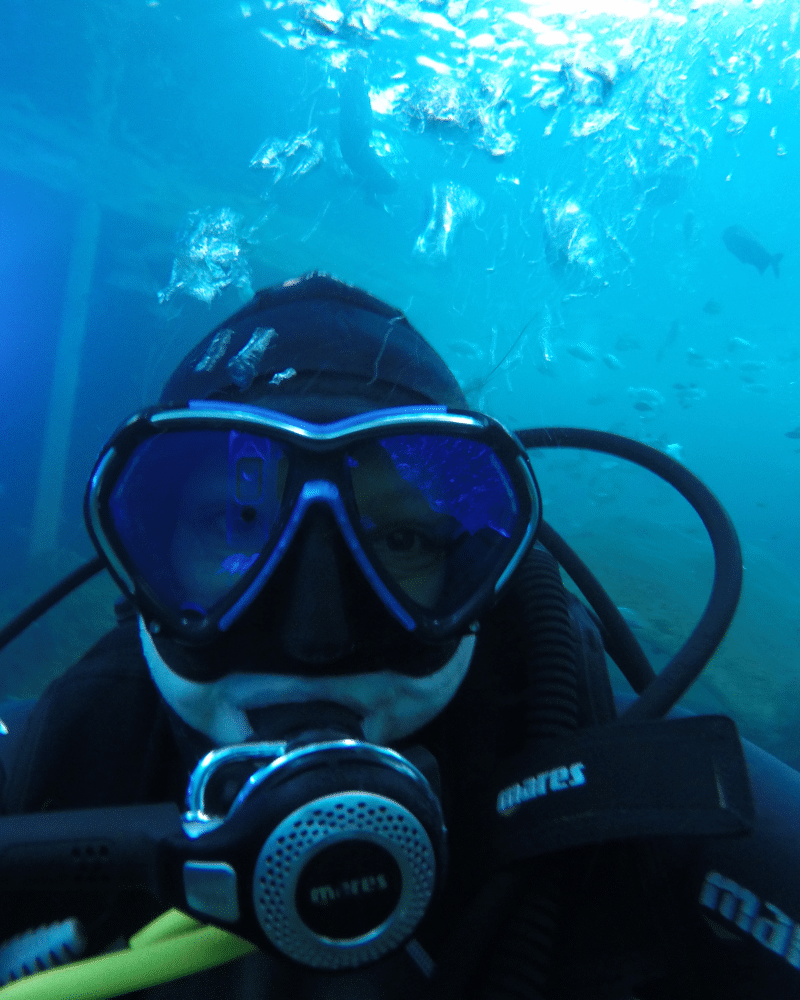 Onderwaterselfie van Heidi in de indoorduiktank van TODI in Limburg. Boven haar stijgen kleine luchtbelletjes op. Ze heeft een zwart duikmasker op en een zwart ademautomaat in haar mond.