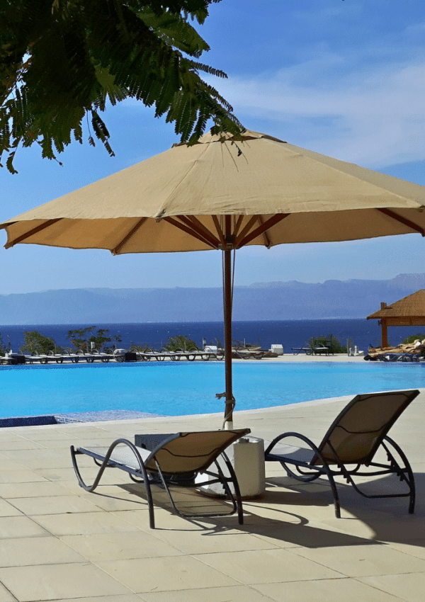 Twee strandstoelen staan onder een parasol, aan de rand van het zwembad van het hotel. In de verte zie je de zee en omliggende heuvels.