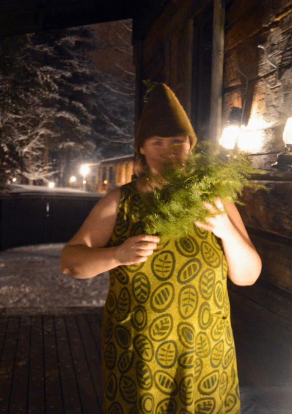 Whisking, met berkenblad, behoort tot de Finse saunatraditie. Op de foto een vrouw met bladeren in haar handen en een typische Finse saunamuts op haar hoofd. Ze draagt een groen kleedje. Het is donker en er ligt sneeuw. Er branden een paar lampjes aan het houten huisje.