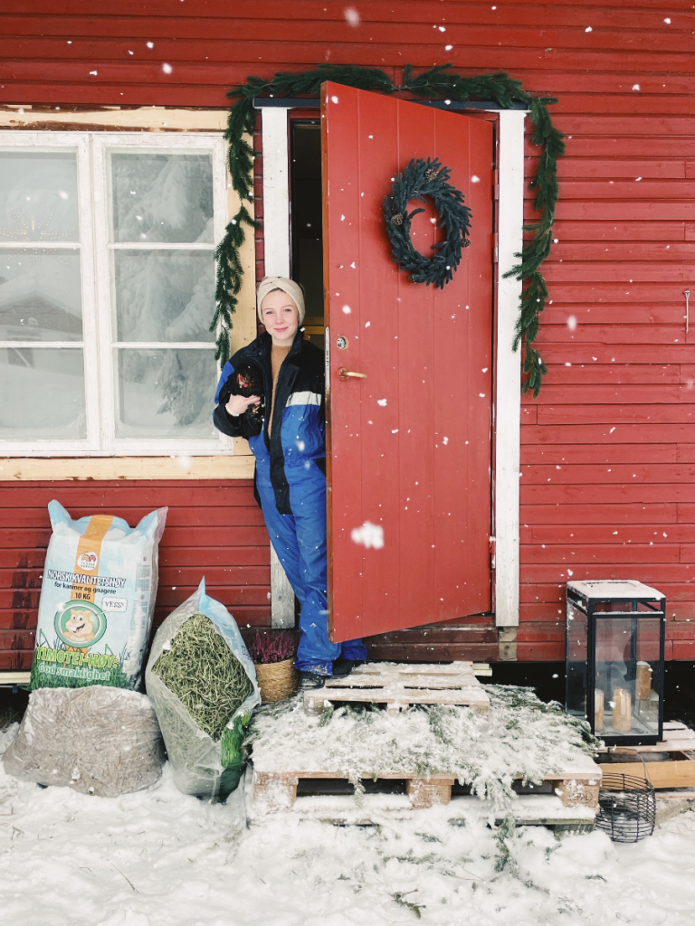 Smiling girl standing in the doorway of a red backyard coop with chicken under her arm. Snow on the ground.