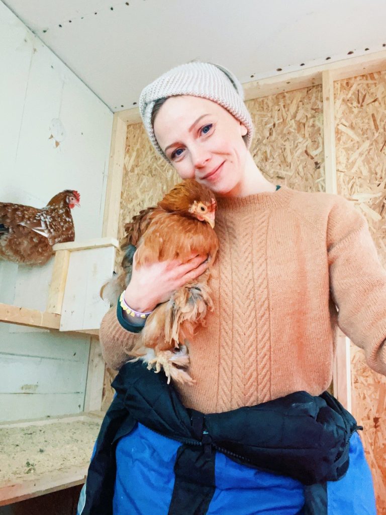 Smiling girl holding a chicken inside a coop. She is wearing a snow suit, wool sweater and headband because it's winter and cold outside. Behind them another hen is roosting.