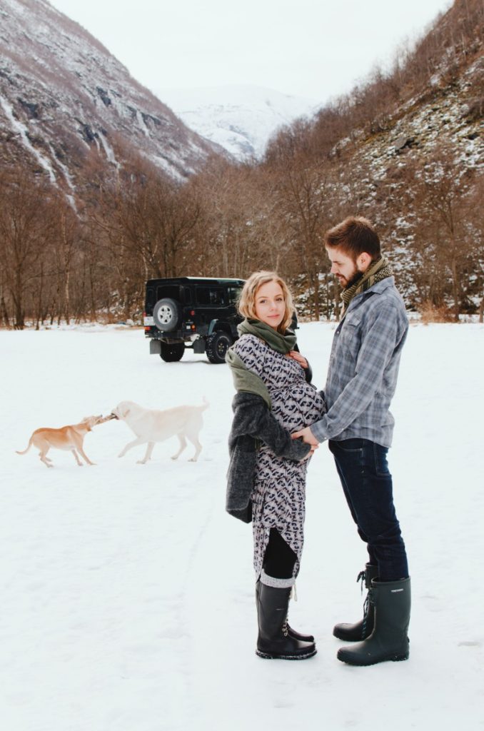 A couple holding hands, standing towards each other. Dogs playing in the background by a parked old land rover defender.