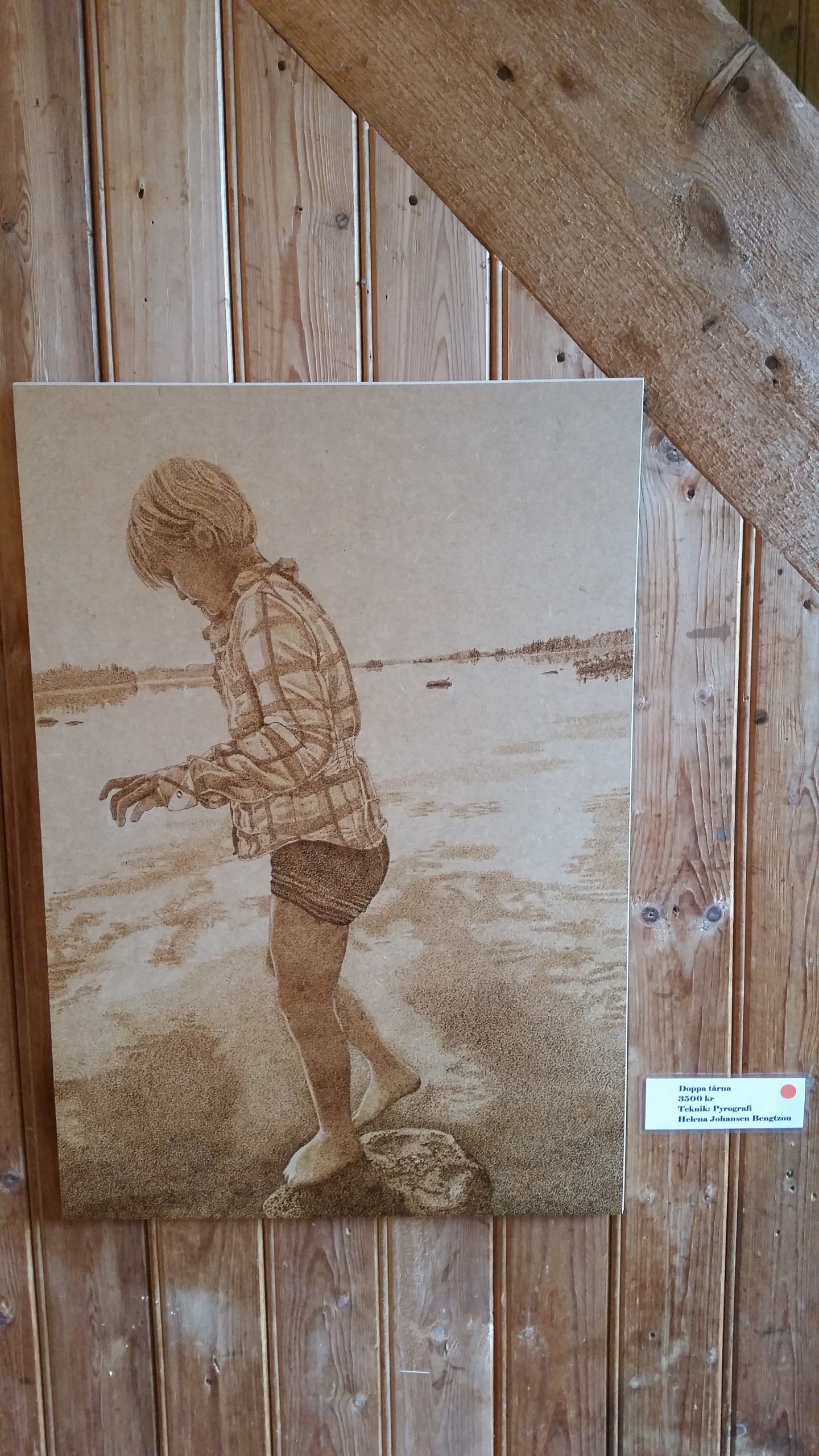 trätavla med glödritat motiv, ung flicka med shorts och skjorta i helfigur vid strand som tar ett steg ut i vattnet