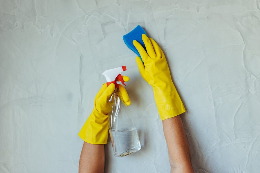 hands-cleaning-wall-with-spray-bottle-and-sponge-28576631 Close-up of hands in yellow gloves cleaning a wall with a spray bottle and sponge.