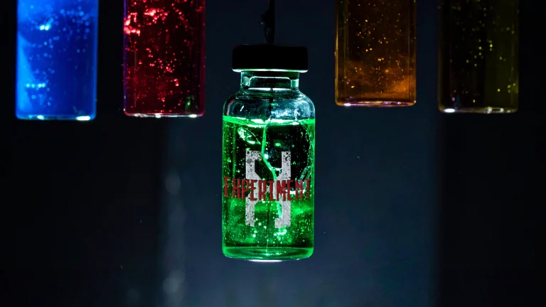Five glowing vials—blue, red, green, orange, yellow—hanging from the ceiling in a dark lab, with the bright green center vial marked 'Experiment X'