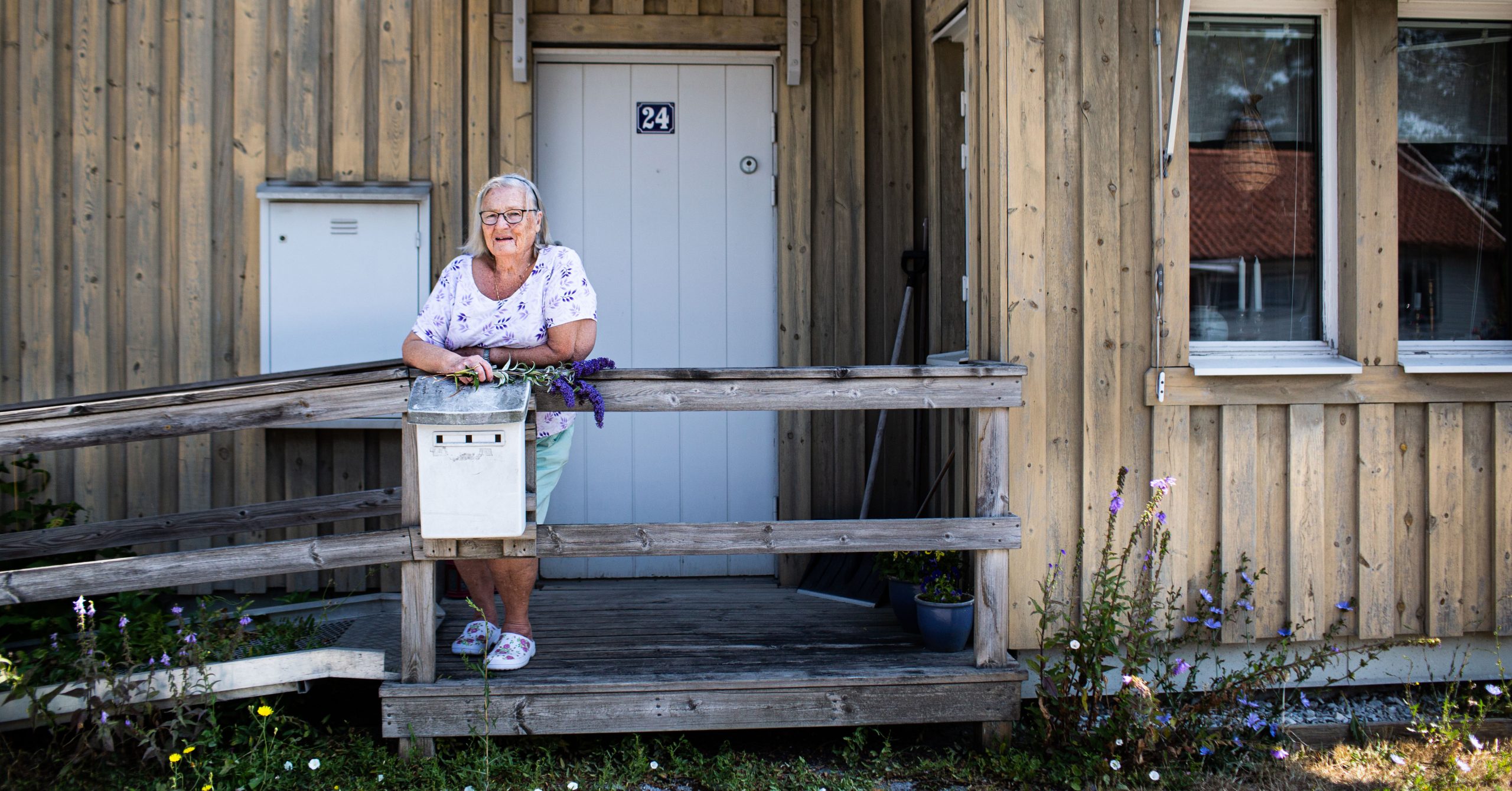 En äldre dam står utanför sin dörr med blommor i handen