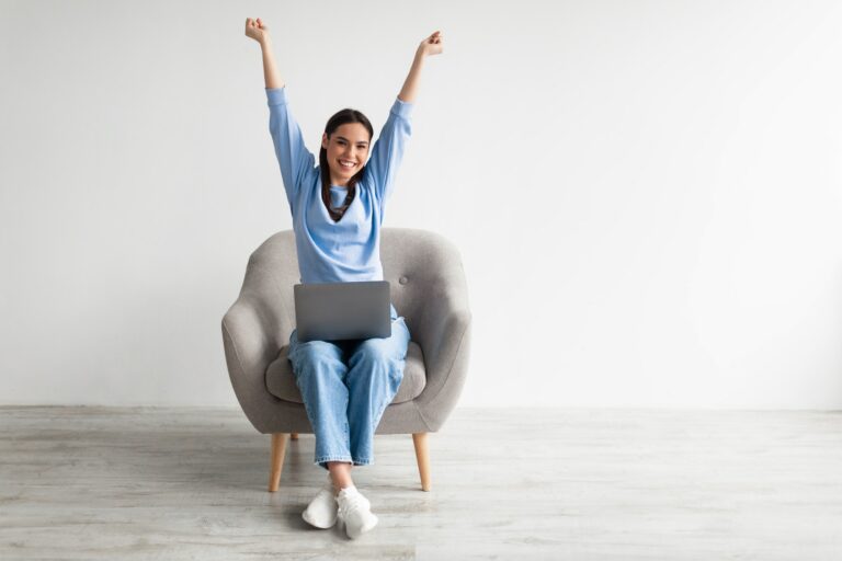 Caucasian woman in armchair raising hands up, celebrating online win, enjoying sale in web store