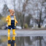 Happy little boy in yellow raincoat jumping in puddle after rain in autumn day.