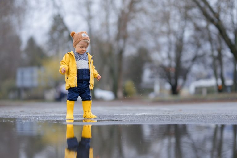 Happy little boy in yellow raincoat jumping in puddle after rain in autumn day.