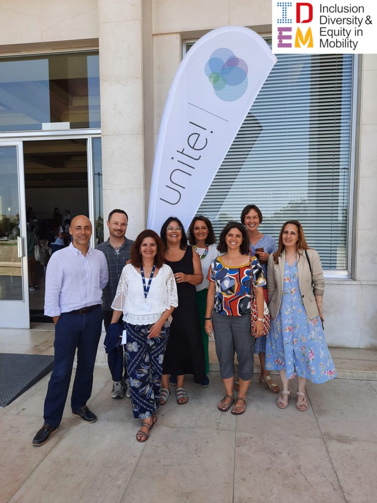 IDEM Team Members are smiling at the camera in front of an IDEM Flag at the Unite! Student Festival in University of Lisbon