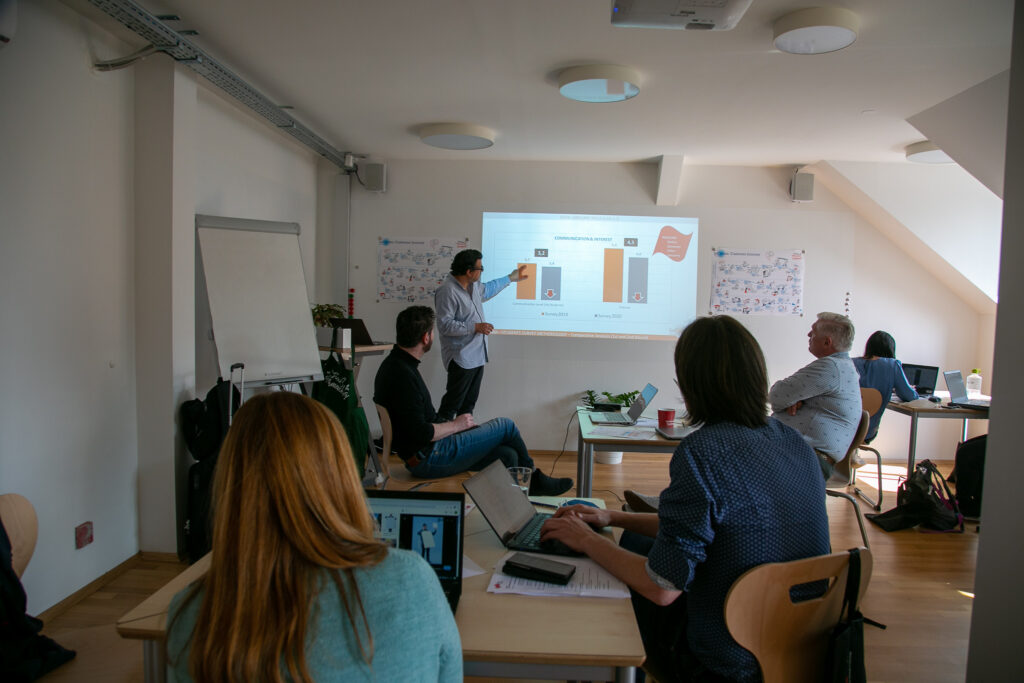 One man presents a powerpoint slide while others listen in a classroom setting.