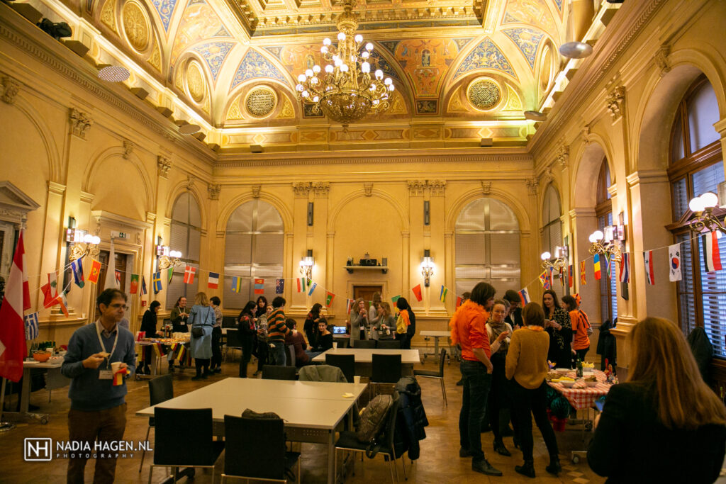 a group of around 30 people socialize in a yellow-lit room with high, painted ceilings and a chandelier