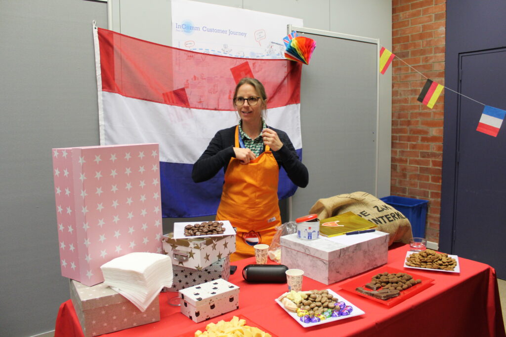 a woman in an orange apron is standing in front of a Dutch flag and in front of a table full with cookies