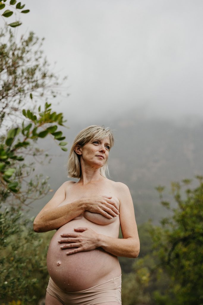 naken gravid kvinne holder hendene på magen under en naturlig gravidfotografering i bergen