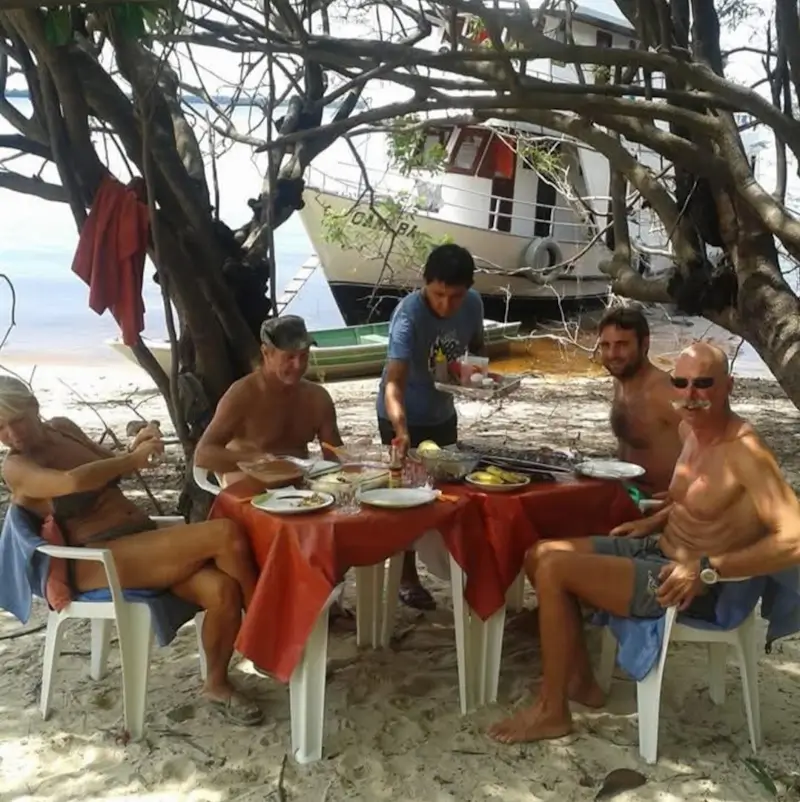 Small group sitting under a tree on an Amazon river beach with a boat in the background