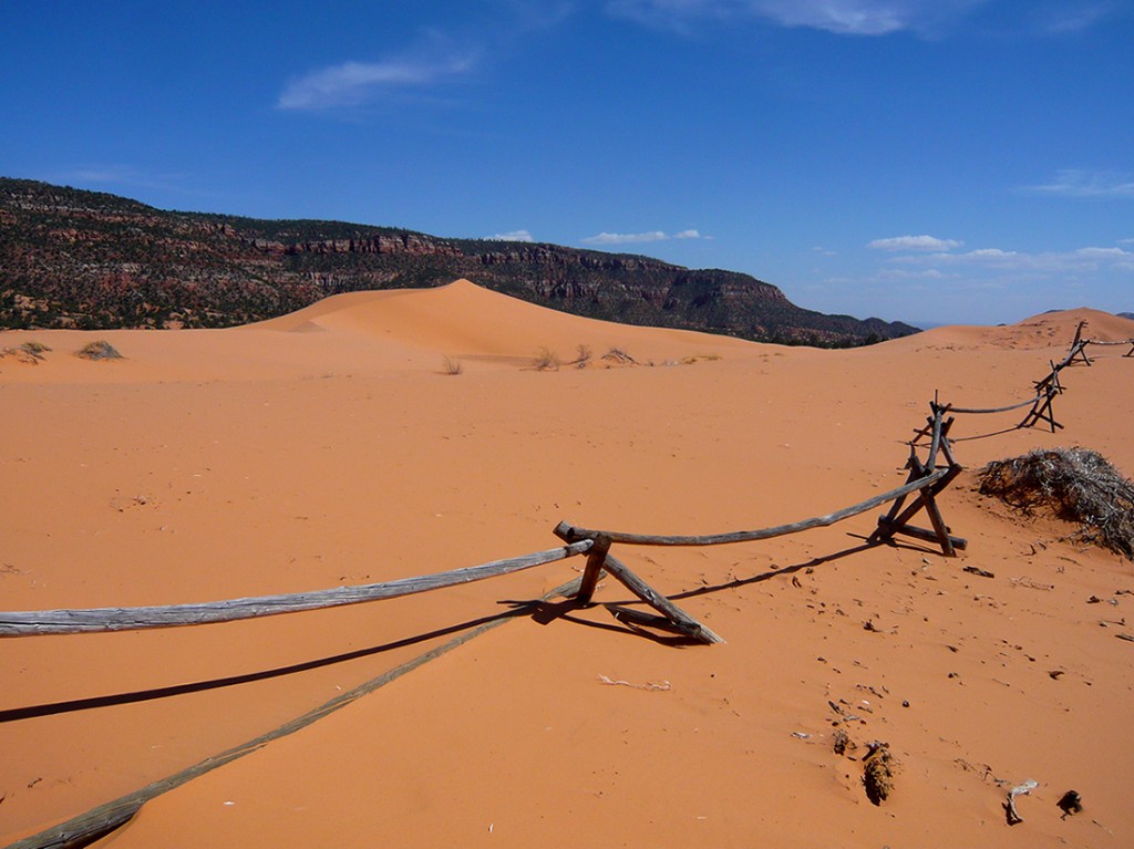 sagging wooden fence running into the distance casting a shadow on pink sand, sand dunes and cliffs in the distance against a blue sky