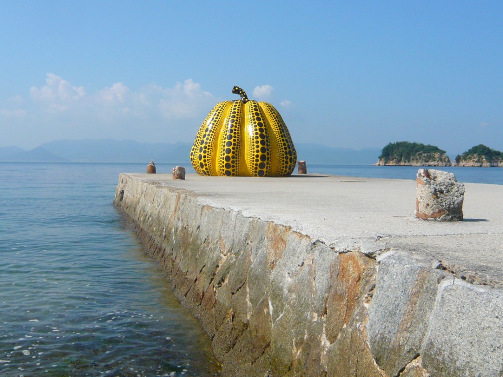 view of a large fiberglass, yellow pumpkin covered in with back dots on a cement pier surrounded by the sea with rocky islands in the background