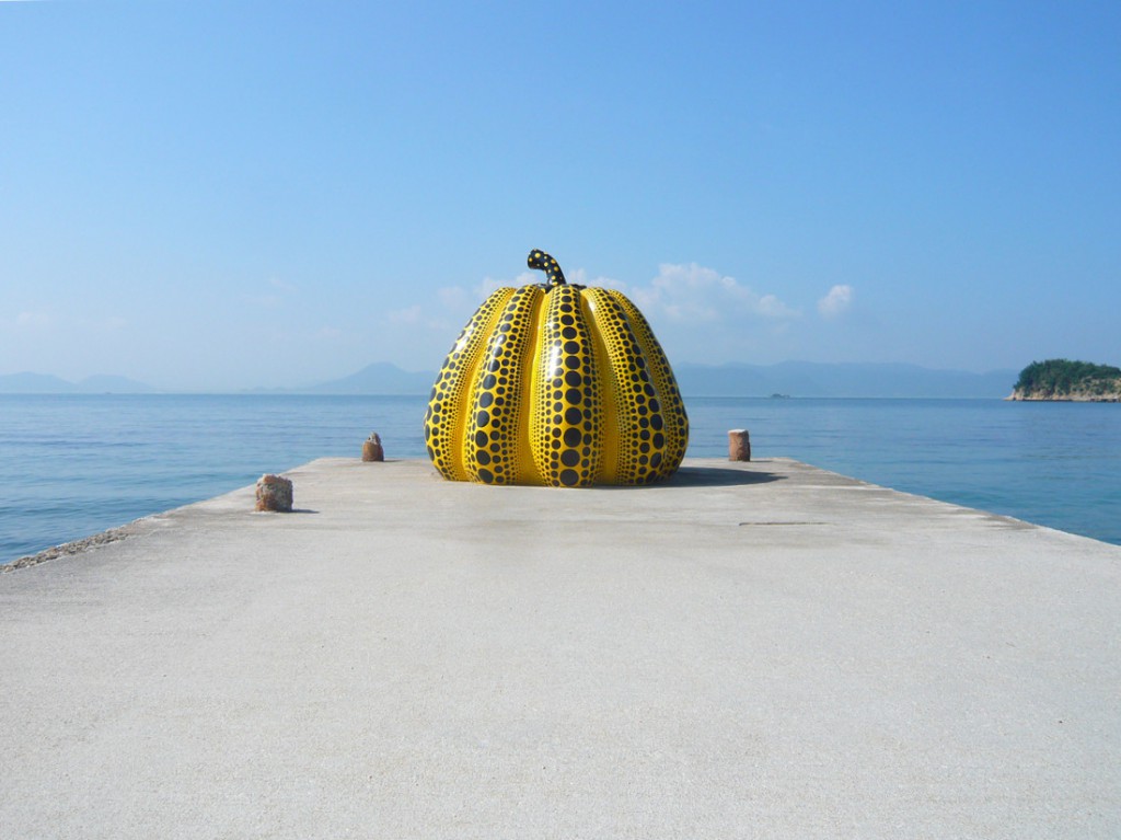 view of a sculpture of a large yellow, fiberglass pumpkin, covered in black spots on a cement pier