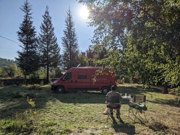 A picture of our red campervan on a campsite in Portugal. It is parked up between trees, and the sky is completely blue, you can see the sun in the middle of the picture. Rene is sat on a camping chair behind a table with tea pot and eating stuff around it. There are lots of trees. We are the only ones there.