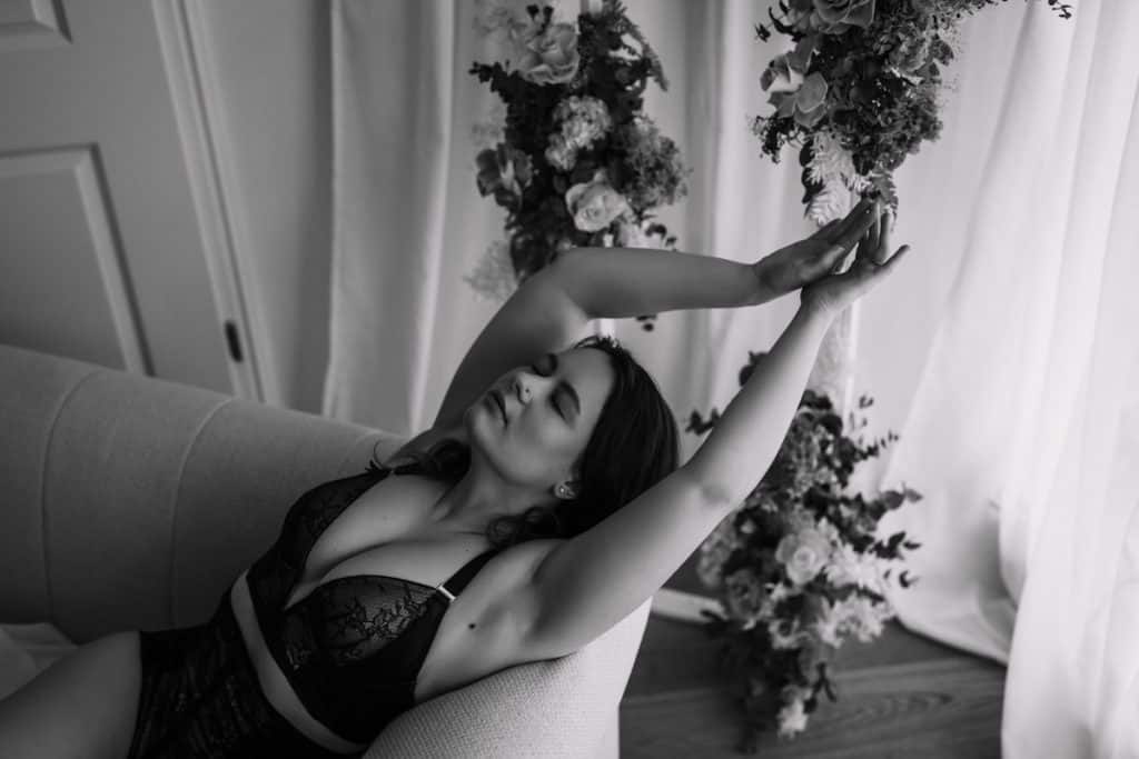 Schwarz-weiß-Fotografie einer Frau, die auf einem Sofa sitzt und ihre Arme entspannt nach oben streckt. Sie trägt schwarze Spitzenunterwäsche und schließt die Augen, während sie in einer ruhigen, eleganten Pose verweilt. Im Hintergrund sind Blumenarrangements und sanft fallende Vorhänge zu sehen, die eine romantische Atmosphäre schaffen.