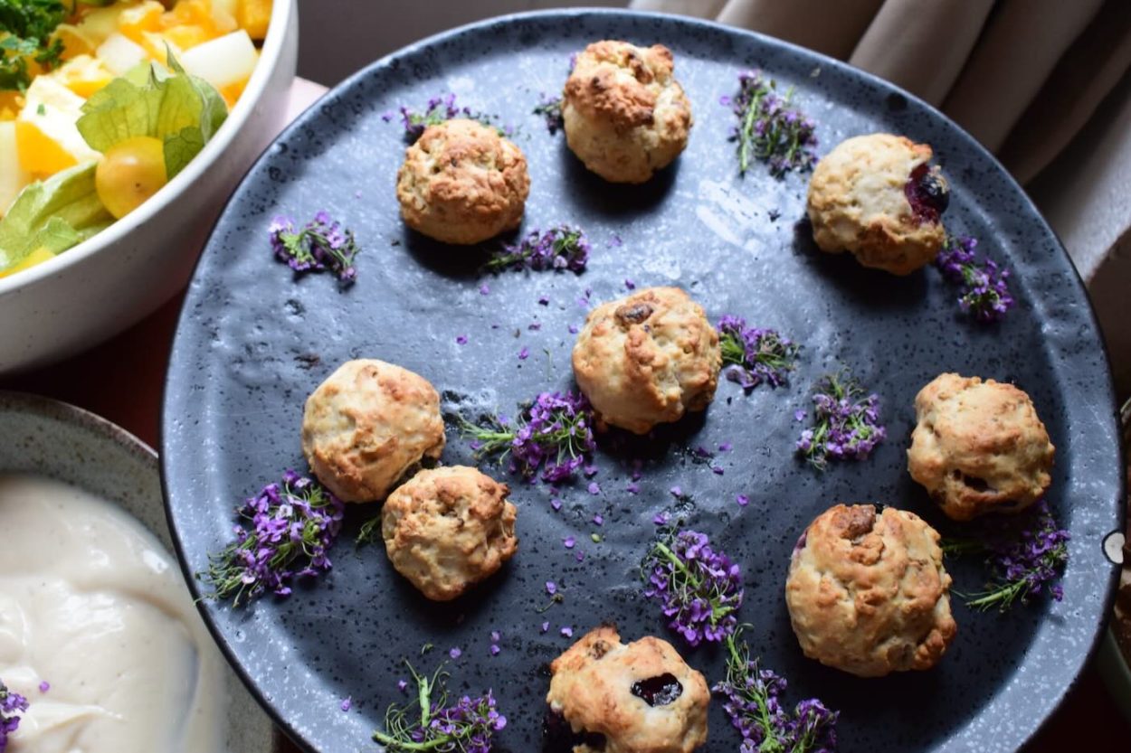 Das Bild zeigt eine kreative Snackvariation aus frisch gebackenen, veganen Scones, liebevoll angerichtet auf einem dunklen Keramikteller und umgeben von dekorativen Lavendelblüten. Dieses feine, pflanzenbasierte Gebäck war Teil des Retreat-Buffets beim Yoga Wochenende „Be Love ♡ Be Light ♡ Be You“ mit JayJay & Hannah im Seminarzentrum Grube Louise. Die Kombination aus Geschmack und Ästhetik macht dieses Bild zu einem visuellen Genuss – und zu einem inspirierenden Einblick in die Retreat-Küche.