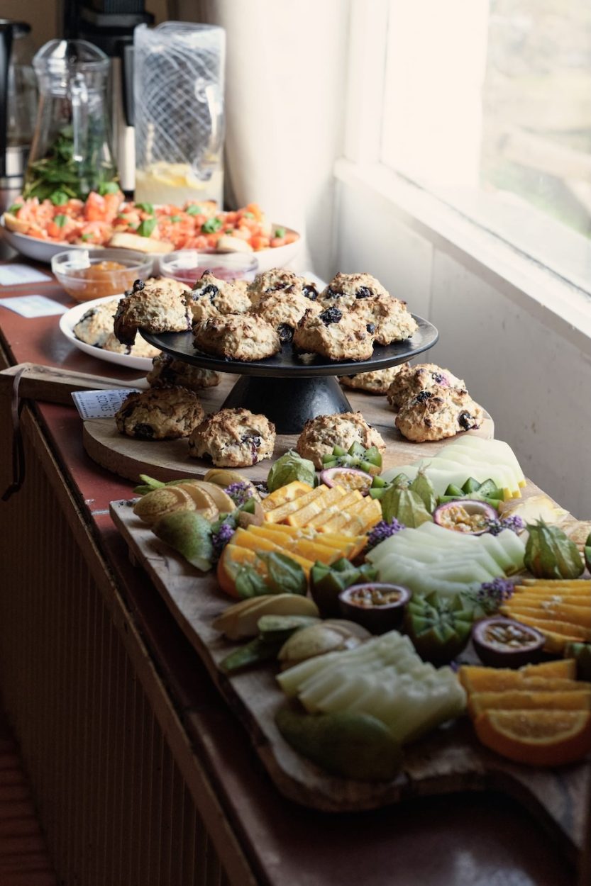 Das Bild zeigt das vegane Buffet beim Yoga Retreat „Be Love ♡ Be Light ♡ Be You“ mit JayJay & Hannah in der Grube Louise im Westerwald. Im Mittelpunkt steht eine reich gedeckte Tafel mit saisonalem Obst wie Mango, Physalis, Birne und Passionsfrucht, begleitet von frischen Scones mit Rosinen. Der natürliche Lichteinfall unterstreicht die warme, einladende Atmosphäre. Das Essen wurde von Nina Joy (@njoysoulfood) mit Liebe zubereitet – als nährende Ergänzung zu einem tief transformierenden Retreat-Wochenende.