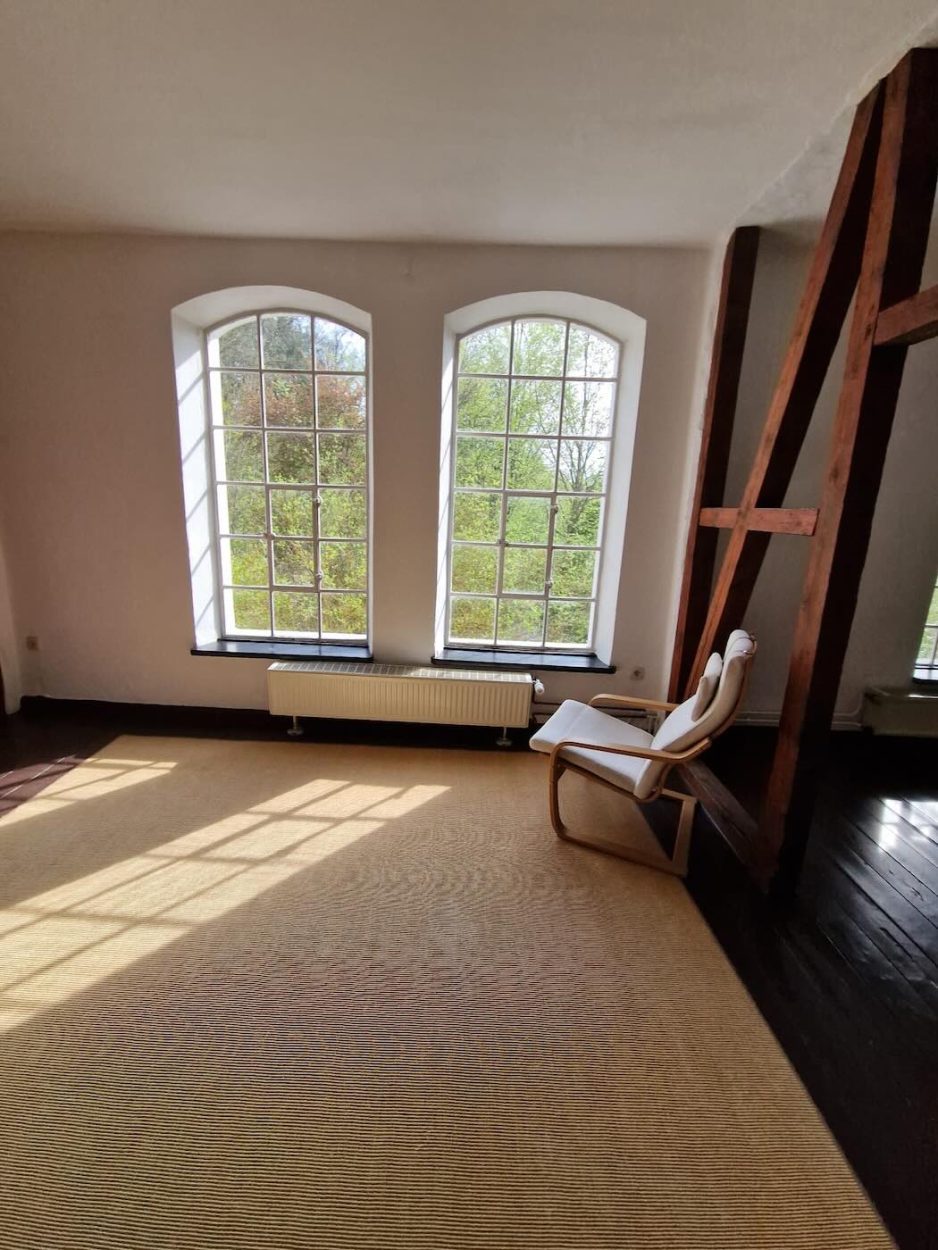 Dieses Bild zeigt den lichtdurchfluteten Yogaraum mit hohen Fenstern im Seminarzentrum Grube Louise im Westerwald. Der ruhige, minimalistisch eingerichtete Raum lädt zur Einkehr ein und bietet ideale Bedingungen für Yoga, Meditation und Atemarbeit im Rahmen des Be Love ♡ Be Light ♡ Be You Retreats mit JayJay Yoga. Das warme Licht und der Blick ins Grüne schaffen eine besondere Atmosphäre der Offenheit und Stille.