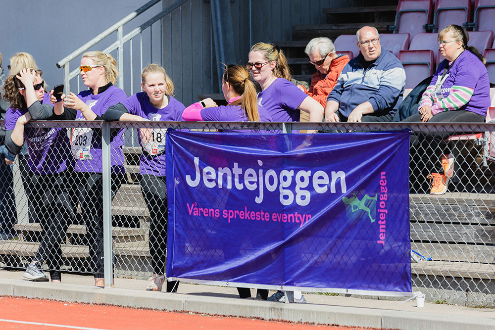 Deltakere og tilskuere som står ved en løpebane bak et "Jentejoggen"-banner