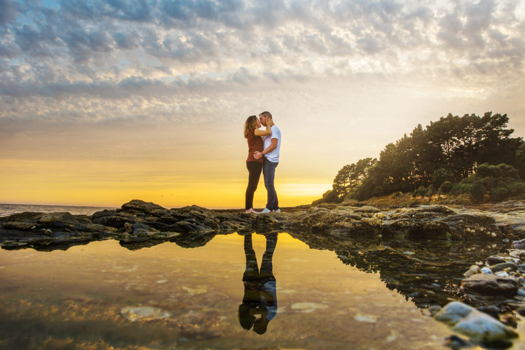 Couple qui s'embrassent sur les rochers avec leur reflet dans l'eau pendant le coucher de soleil