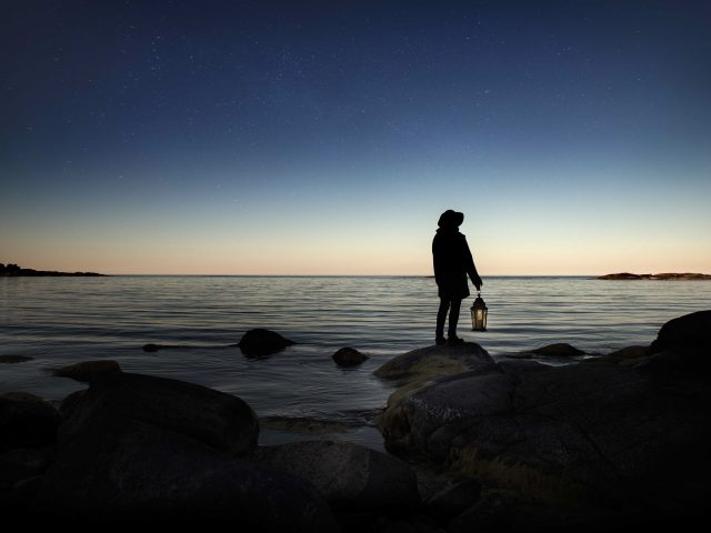 Person med brinnande lykta står vid ett hav och spejar ut i natten