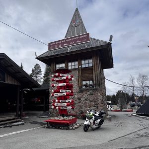 Holzgeb&auml;ude mit spitzem Turm, daneben Stra&szlig;enschild mit vielen Richtungen und ein geparktes Motorrad, bew&ouml;lkter Himmel.
