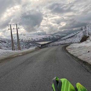 Verschneite Straße mit Bergen im Hintergrund, Strommasten am Straßenrand, bewölkter Himmel, ein grünes Objekt im Vordergrund.
