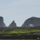 Steile Klippen am Strand; eine einsame Person läuft entlang während sich eine große Welle an einer Klippe bricht. Im Vordergrund sieht man gelbe Blumen.