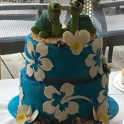 Eine zweistöckige Hochzeitstorte im Hawaii-Look. Die blaue Torte ist mit weißen Hibiskusblüten und Orchideen verziert und ganz oben ist eine Marzipanfigur, zwei Schildkröten, die ein Hochzeitspaar darstellen.