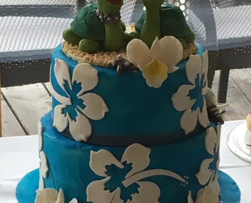 Eine zweistöckige Hochzeitstorte im Hawaii-Look. Die blaue Torte ist mit weißen Hibiskusblüten und Orchideen verziert und ganz oben ist eine Marzipanfigur, zwei Schildkröten, die ein Hochzeitspaar darstellen.